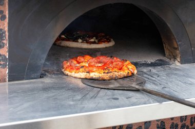 Hareketli bir mutfakta, altın-kahverengi bir pizza bir tuğla fırının sıcaklığından kaldırılır, peyniri köpürür ve malzemeler parlarken, diğer bir pasta sabırla servis edilmek üzere arkasında bekler..