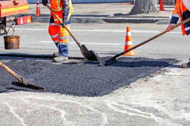 İnşaat işçileri, açık mavi gökyüzünün altındaki bir şehir yoluna özenle yeni bir asfalt seriyorlar. Turuncu güvenlik konileri bölgeyi kuşatarak araçlar için güvenli geçiş sağlıyor..
