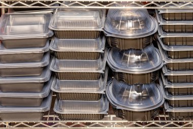 Sağlam siyah yiyecek kapları metal raflarda üst üste istiflenir, ambalaj ve teslimat için hazırdır. Temiz kapakları içerideki yemekleri gösteriyor. Kalabalık bir mutfakta organize olmuş kaosu gösteriyor..