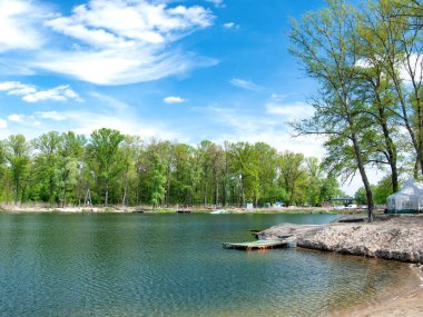 Huzurlu bir göl manzarası, sahili çevreleyen canlı yeşillik ve açık mavi gökyüzü ile doğanın güzelliğini yakalar. Sakin su, huzurlu çevreyi yansıtır. Rahatlamayı davet eder..
