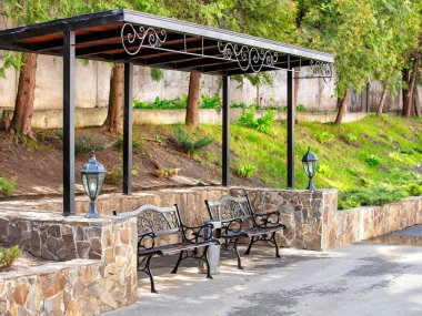 Two ornate metal benches rest beneath a beautifully designed canopy, surrounded by lush greenery. Soft sunlight filters through trees, creating a peaceful atmosphere for visitors. clipart