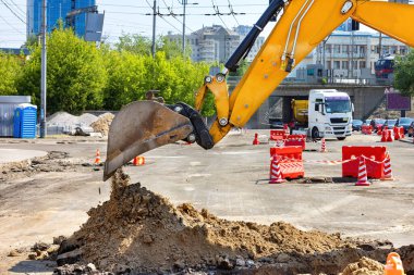 A yellow excavator digs into the ground, lifting a mound of earth on a busy urban construction site. Construction vehicles and safety barriers can be seen in the background under a bright sky. clipart