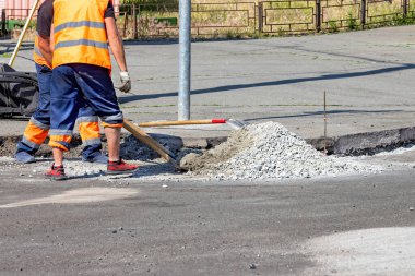 Parlak yelekli inşaat işçileri bir şehir caddesini onarmakla, açık mavi gökyüzünün altındaki kaldırıma taze beton sürmekle, takım çalışması ve adanmışlık sergilemekle meşguller. Boşluğu kopyala.