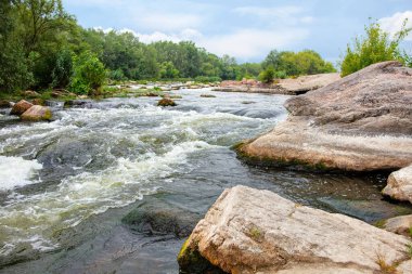 Bereketli yeşilliklerle çevrili sakin bir nehir pürüzsüz taşların üzerinden akıyor. Doğanın durgunluğu bu bulutlu günde barışçıl bir atmosferi çağrıştırıyor, yansımayı ve bağlantıyı davet ediyor..