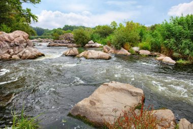 Akan sular, canlı yeşillikler ve sakin bir atmosferle çevrili pürüzsüz taşların etrafında yumuşak dalgalar oluşturur. Gökyüzü yağmuru işaret ediyor, doğanın renklerini güzelce harmanlıyor..