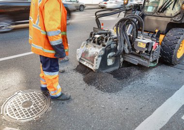 Parlak turuncu teçhizatlı bir inşaat işçisi kaldırımı onarmak için güçlü bir makine kullanıyor. Yoğun kentsel ortamı ve yol bakımının aciliyetini vurgulayan arabalar yakınlaştırılıyor.