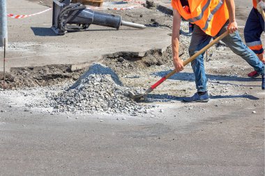 Güvenlik yelekleri giyen iki inşaat işçisi yol onarım projesinde sıkı çalışıyor. Biri çakılları küreklerken diğeri yakınlarda yardım ediyor. Parlak, güneşli bir gün. Boşluğu kopyala.