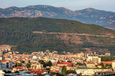 Güneş görkemli dağların ardında batarken, nefes kesici renkler gökyüzünü kaplıyor. Aşağıdaki sakin Budva şehrine altın bir parıltı saçıyor. Evler ve binalar sessiz sokaklarda çeşitli mimari yapılar sergiliyor..