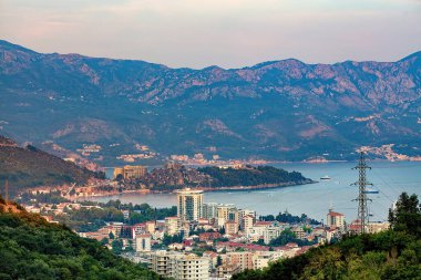 Sakin Budva kıyı kasabası, alacakaranlık çökerken görkemli dağlarla çevrili kıyı şeridini kucaklıyor. Ufuk çizgisi, doğa ve şehir yaşamını uyum içinde harmanlayan modern binalarla dolu..
