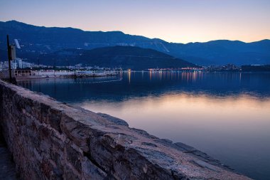 Budva limanına alacakaranlık inerken sakin sular kıyıları kaplıyor. Komşu kasabanın yumuşak ışıkları yüzeyde parıldıyor ve bu resim gibi yerde sakin bir atmosfer yaratıyor..