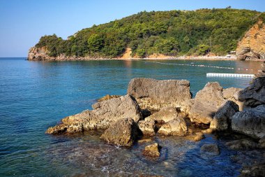 Dalgalar, Budva 'daki Mogren Sahili' nin sakin kıyı şeridi boyunca kayalara nazikçe çarpıyor. Bereketli yeşilliğin berrak mavi denizle buluştuğu yerde. Güneş ışığı suda parlıyor, sakin bir yaz atmosferi yaratıyor..