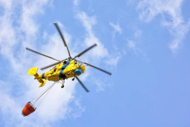 Parlak sarı bir helikopter mavi gökyüzünün zeminine doğru süzülüyor. Suyu ustalıkla taşımak için manevra yapıyor..