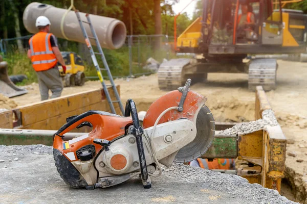 Orange petrol saw with a diamond blade for cutting concrete against a blurred background of drainage works on construction site