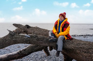 Parlak sarı ceketli bir kadın soğuk kış gününde deniz kenarında tek başına yürüyor. Seyahat yaşam tarzı konsepti
