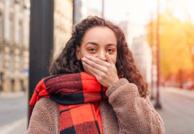 Ceketli, kıvırcık saçlı ve elini dudaklarına dayayan turuncu ceketli bir kadın. Sessizlik altın bir kavram.