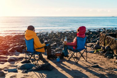 Kışın plajda plaj sandalyelerinde iki kişi, güneşli, mavi gökyüzü ufkunda. İngiliz soğuk kışı. Yerel turizm konsepti.