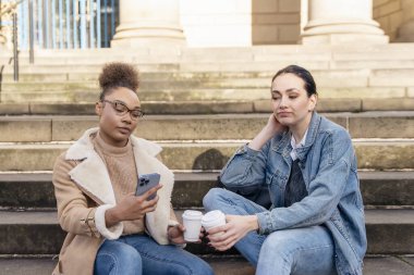 Kot ceketli iki kız öğrenci telefonda birbirleriyle konuşuyor, gülüyorlar ve şehirdeki merdivenlerde kahve içiyorlar. dijital bölünme kavramı