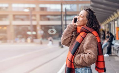 Ceketli ve turuncu eşarplı genç bir kadın durakta tramvay bekliyor ve pofuduk hayat tarzı fotoğrafı hakkında konuşuyor.