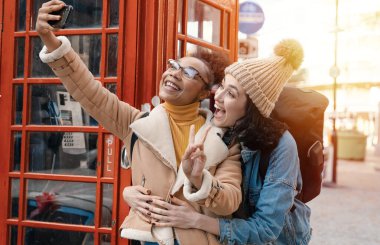 two friend, girlfriend and women using a mobile phone, camera and taking selfie against a red phonebox in the city of England.Travel Lifestyle concept