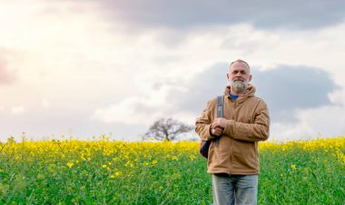 Sakallı, mutlu, kolza tohumu tarlasında sırt çantalı bir emekli. Seyahat yaşam tarzı konsepti