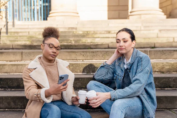 two student women in denim jacket is talking to each other, on the phone, laughing, and drinking coffee on stairs in city.  digital divide concept
