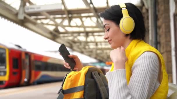 在火车站用耳机听音乐的年轻女子带着音乐欣赏旅行的概念旅行 — 图库视频影像