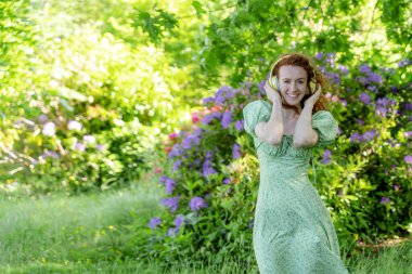 Açık havada, yeşil elbiseli ve kulaklıklı, halka açık parkta dans eden mutlu kızıl saçlı kadının portresi. Çevrimiçi müzik dinleyip kendin ol.