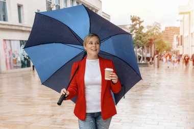 Kırmızı ceketli bir kadın şemsiyesinin altında kahve ile modern bir şehirde dolaşıyor.