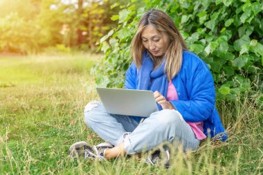 Mavi ceketli Asyalı kadın halka açık parkta dizüstü bilgisayar kullanıyor. Her yerde internet kullanıyor. Yaşam tarzı kavramı.