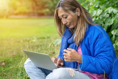 Mavi ceketli Asyalı kadın halka açık parkta dizüstü bilgisayar kullanıyor. Her yerde internet kullanıyor. Yaşam tarzı kavramı.