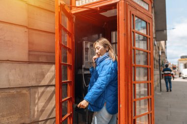 Mavi ceketli Asyalı kadın İngiliz şehrinde yürüyor ve kırmızı kutuda telefonla arıyor.