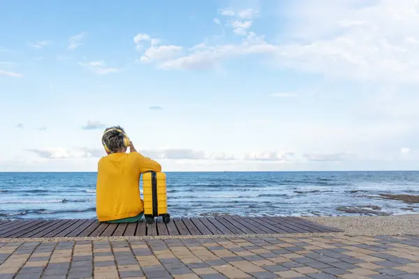 Woman in a yellow jacket with a yellow suitcase and headphones on the seaside on hot sunny day arrived in a tourist town. Travel Lifestyle concept
