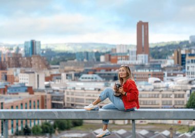 Kırmızı ceketli mutlu Asyalı kadın Sheffield 'ın önünde oturuyor ve harika bir yaz gününde fotoğraf çekiyor.