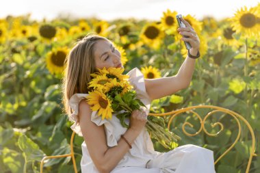 Beyaz elbiseli, buketli neşeli Asyalı kadın Yorkshire 'da ayçiçeği tarlasında selfie çekiyor.