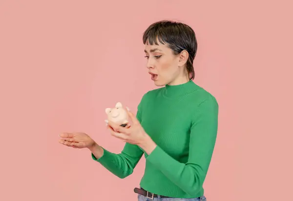 Sad woman wearing green casual sweater discovering empty piggy bank without savings. Investment, saving money, currency, deposit. Indoor studio shot isolated on pink background.