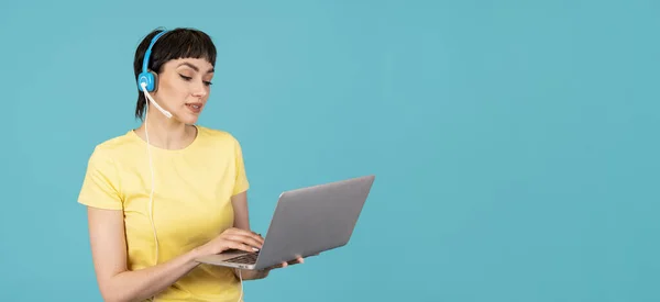 Amazing woman in a yellow t-shirt and headphones using laptop, pc computer on blue background with copy space. Work studying, virtual training, e-learning, watching online education webinar concept