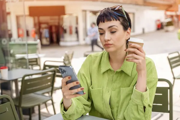 thoughtful woman  in green jacket having coffee at the street cafe outside using phone, having fun time. lifestyle concept