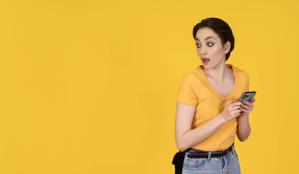 surprised woman in yellow t-shirt and jeans using a mobile phone on yellow background with copy space