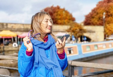 Mavi ceketli ve gözlüklü mutlu Asyalı kadın rıhtımda yürüyor, bankta oturuyor, yata, yelkenliye, tekneye bakıyor, telefonda yaşam tarzı seyahat konseptiyle konuşuyor.