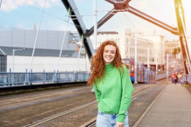 Yeşil kazaklı, dişleri açık gülümseyen ve şehir caddesinde bulanık tramvayın arka planında yürüyen genç, kızıl saçlı bir kadın. Mutlu Yaşam Tarzı konsepti