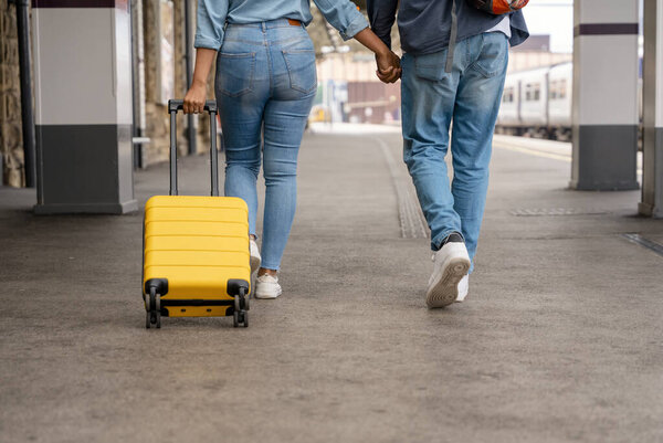 Счастливая любящая пара с багажом и рюкзаком прогуливаясь вдоль железнодорожной платформы, как они готовы путешествовать в отпуск. Мужчина и женщина держатся за руки, оглядываются назад и улыбаются. Путешествие на поезде