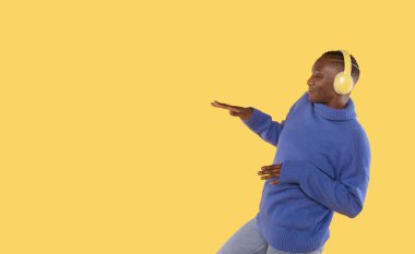 Sarı arka plan stüdyo resminde izole edilmiş mavi kazaklı neşeli genç kadın. İnsanların yaşam tarzı konsepti. Kopyalama alanı oluşturun. Kulaklıkla müzik dinlemek, dans etmek