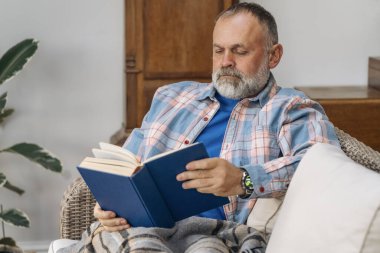 Gülümseyen yaşlı, olgun bir adam gülüyor kitap okuyor, dinleniyor, oturma odasında kanepede oturuyor.