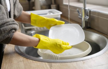 Sarı eldivenli kadın ev mutfağında bulaşık yıkıyor. Güzel genç ev kadınlarının ev işlerini yapmasının yan görüntüsü. Kadın yıkama tabakları