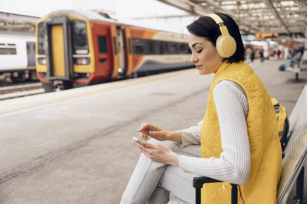 Young woman listening to music via headphones at railway station Travel with music Enjoying travel concept