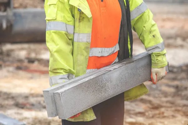 groundworker in orange and yellow hi-viz carrying heavy concrete edging curbs on the building site during new road construction. Manual handling safety concept
