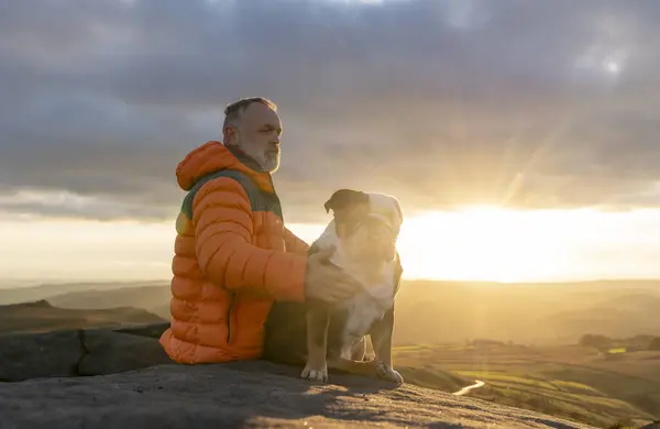 old mature man hiking in mountains with his dog, exercising and fitness for wellness, healthy lifestyle, and smile. Face of a senior mature gentleman with bulldog sitting on a rock, enjoying calm day