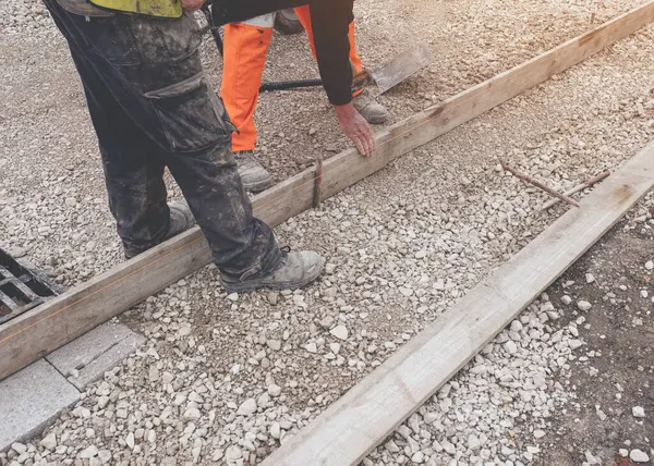 Groundworker in orange safety hi-vis trousers fixing a timber along string line with a steel pin to form a kerb riser and a straight edge for tarmac road surface during new road construction