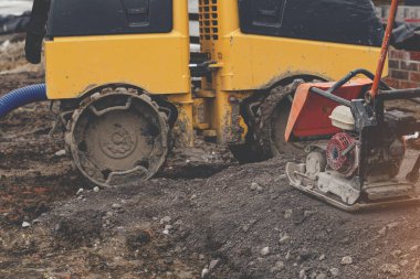 Construction worker using petrol powered plate compactor to compact stone around foundation of new build residential house in front of remote controlled trench compactor