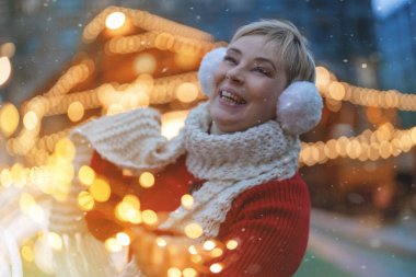 Gülümseyen mutlu kadın şehir merkezinde Noel ışıklarını kullanarak Noel pazarında eğleniyor..